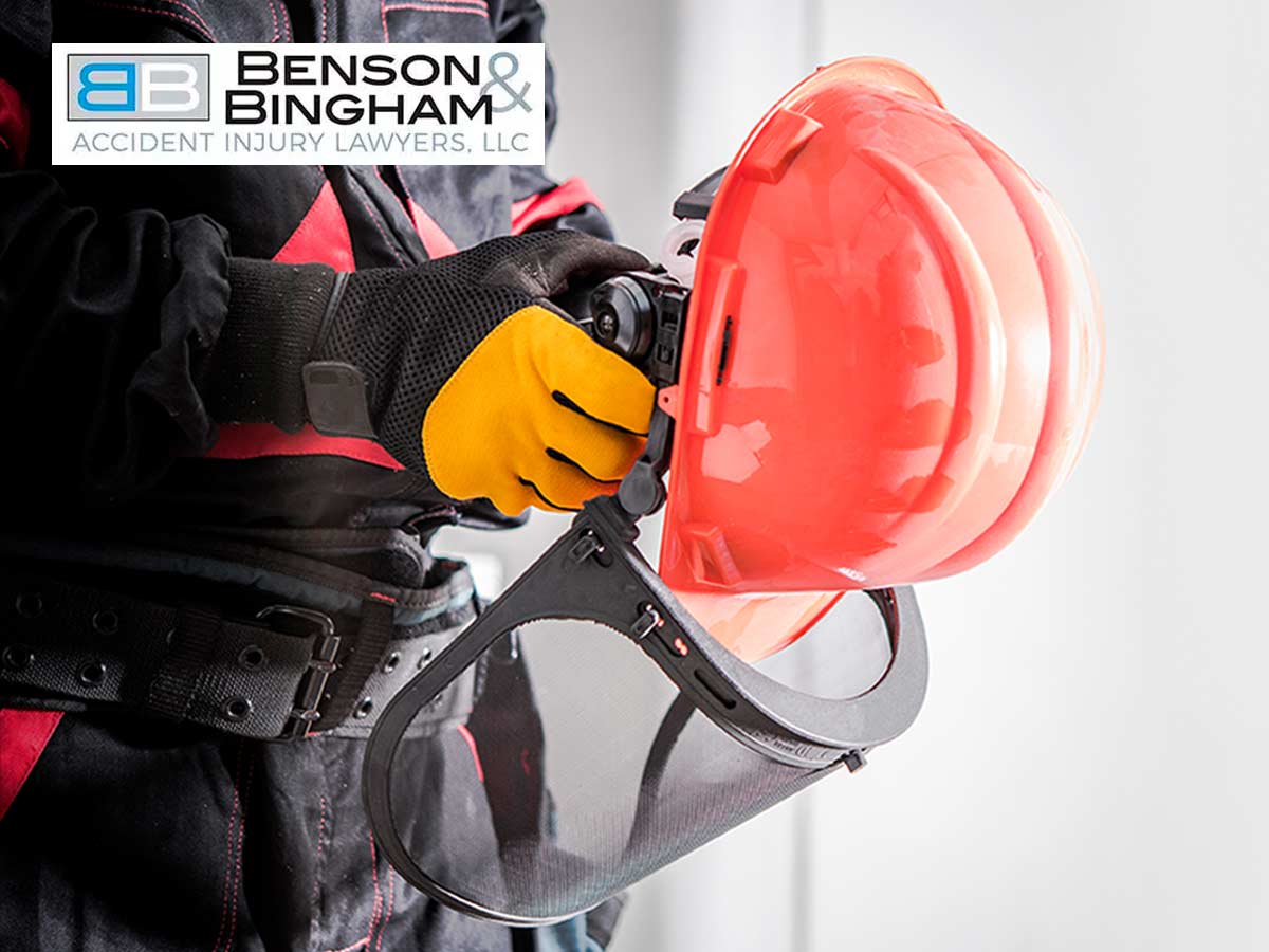A worker in a black and red jacket wearing yellow gloves, adjusting a red hard hat with a face shield, showcasing Personal Protective Equipment